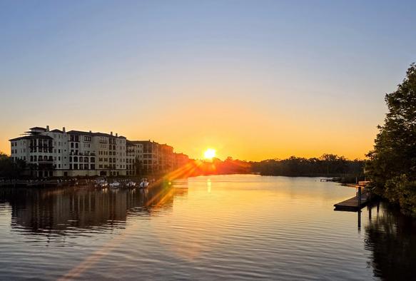 Bright, warm, yellow glowing sunrise over a calm blue waterway with luxurious buildings on one shore and a fishing pier on the opposite side, amongst tropical foliage.