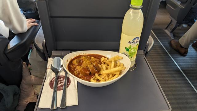 A plate with curry sausage, fries and a bottle of lemonade on the table of a Velaro seat.