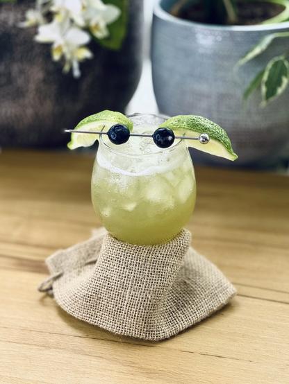 light green cocktail garnished with two lime wedges and two speared blueberries arranged to look like baby Yoda Grogu with a burlap sack wrapped around the glass stem to make it look like a body