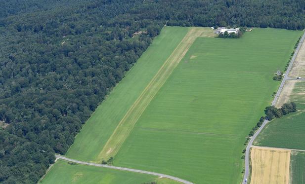 Luftbild des Segelfluggeländes Vielbrunn
Es ist eine grüne Wiese mit Rollsteifen zu erkennen, dahinter befindet sich das Gebäude des Segelflugvereins. Links und hinten wird das Gelände von Wald begrenzt.
Rechts verläuft die Landstraße. Daneben steht der rekonstruierte Wachturm.
(Bild: CC-BY-SA 4.0, 2024, Carsten Steger)