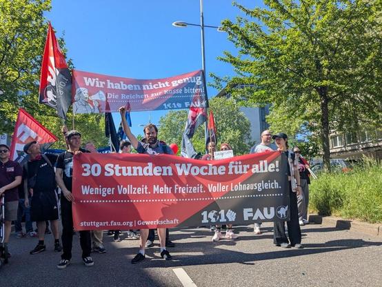Das Foto zeigt das Transparent der FAU Stuttgart mit dem Text: "30 Stunden Woche für alle! - weniger Vollzeit - mehr Freizeit - Voller Lohnausgleich" Dahinter das Hochtransparent mit dem Text: "Wir haben nicht genug - die Reichen zur Kasse bitten!"