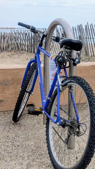 Photo d'un vélo bleu, garé derrière une palissade et le bord de mer. Il a un tract pour le baromètre vélo accroché sur le cadre.