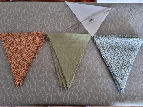 three piles of triangular pieces of fabrics. One in yellow with red stars, one calm green, and one white with blue and yellow flowers on. Above the fabrics is a template in paper and a ruler.