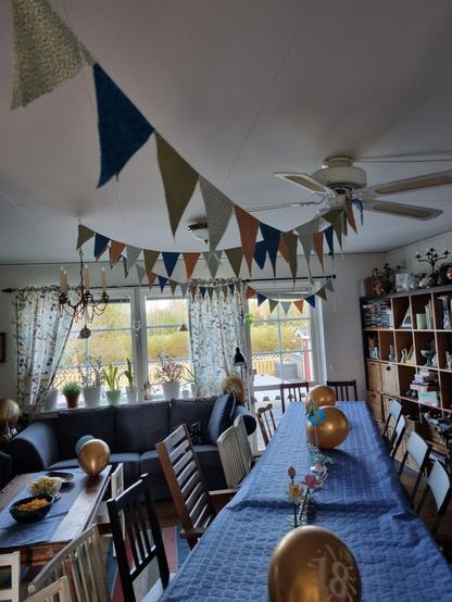 A festive decorated livingroom with a blue table with balloons and serpentines on it and above the pennant garland going in a zig zag pattern from the door opening, to the ceiling fan, to a chandelier to the back of the room.