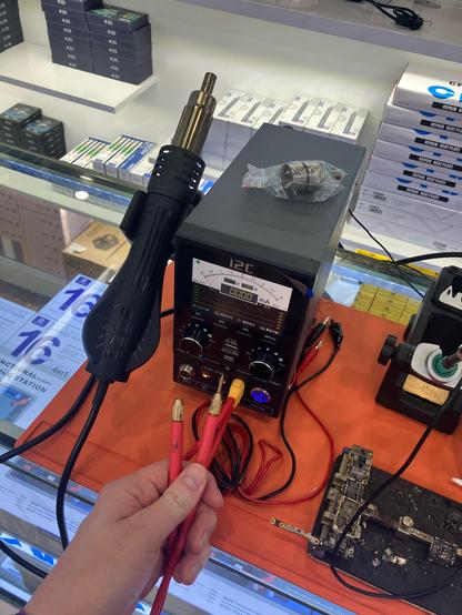 A photo of a small lab bench device with a big display and two turn knobs in the front. To its left a heat gun is mounted, to its right a soldering iron is placed. Coming from the front are two cables that seem to be a spot welder. There are also connectors that hint that the device is a lab power supply. The display shows an analog needle scale and the manufacturer seems to be “I2C”. There are tools in the background