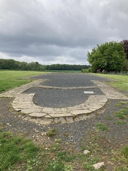 Zu sehen ist auf dem Foto eine Landschaft mit einer archäologischen Erinnerung, die im Boden eingelassen ist.  Es sind im Rund angeordnete helle Natursteine zu sehen, die die Apsis einer frühmittelalterlichen Kirche beschreiben und in das Längsschiff zum Horizont hin verlaufen. Dazwischen ist schwarzer Kies aufgefüllt worden. Der Himmel ist stark bewölkt und den Horizont zieren dunkelgrüne Wälder.