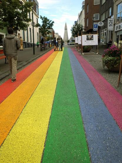 Looking along a street painted like a rainbow red, orange, yellow, green, blue, violet in long stripes- with buildings on both sides and the tower of the  Hallgrimskirkja Church in the middle of frame and in the distance
Reykjavik 2016