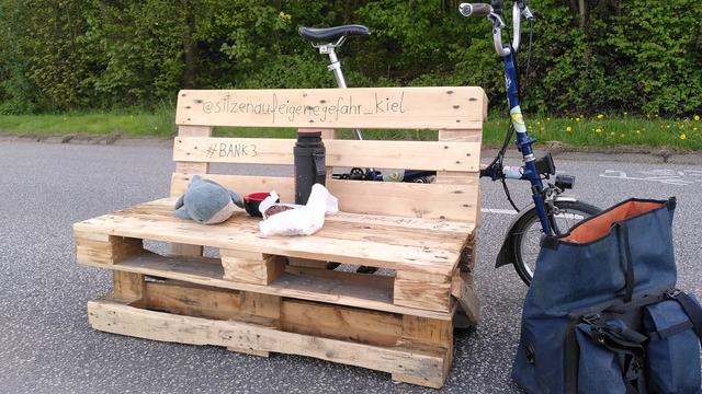 Sitzbank aus Europallette auf einer Straße, Radwegpiktogramm im Hintergrund, dahinter bisschen Rasen und dann dichtes grünes Gebüsch. Rakete, das blaue Faltrad, lehnt von hinten dran, Tasche steht daneben, Blåhaj plushie passt auf Tee aus der Thermoskanne und Nussschnecke auf.
