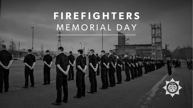 Scottish Fire And Rescue Firefighters memorial day poster. Black and white photograph. Two ranks of Scottish firefighters in dress uniform on the drill ground with fire station and training tower in the distance to right of frame. The backdrop is industrial with power transmission towers. The picture is taken from behind, the firefighters stand at ease, hands clasped behind their backs.