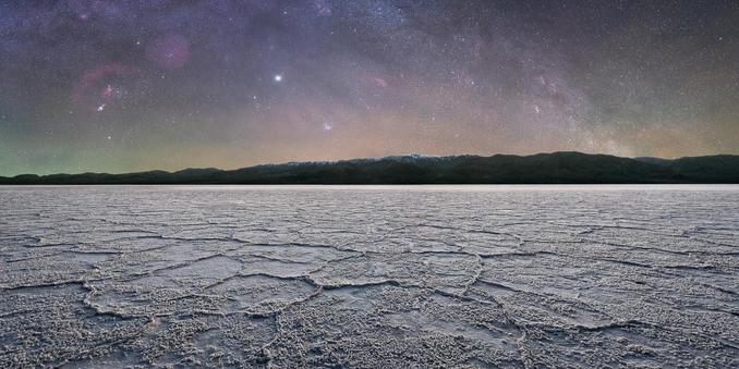Badwater Basin at night. The foreground is the salt flat stretching from your feet toward the horizon. There are ridges forming large, vaguely hexagonal shapes, but there are salt protrusions everywhere. The salt flat is white, but it is nighttime, so the colors are muted. Beyond the salt flat, mountains rise in the distance. The mountains don’t have any peaks, though. 

The sky is the star of the show, pun intended. You can see an arch of stars stretching from side to side. This isn’t the Milky Way as is usually shown because you can’t see the core, but it is the Milky Way all the same. There are stars everywhere. There are varying colors in the sky from air glow. Large reddish “clouds” are throughout the sky, and very notably on the left side. One of the clouds is a crescent that surrounds a circle, centered in the Orion Constellation. This is Barnard’s Loop. Anyway, these clouds are really hydrogen gas structures that the human eye cannot easily see, but by careful equipment choices and post-processing are visible in the photograph.