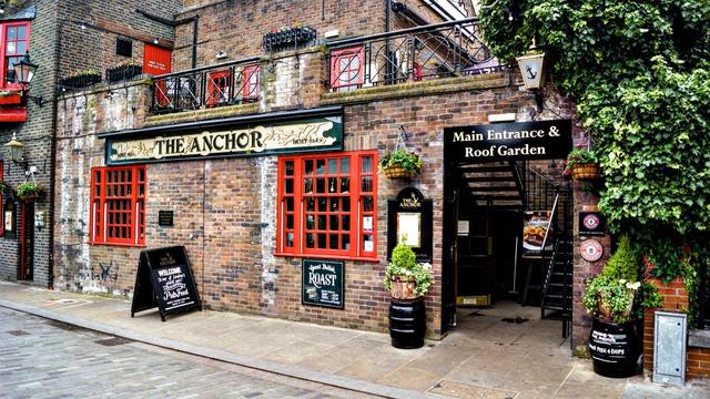 This image shows the exterior of a British pub called "The Anchor." The pub features a classic brick facade with red window frames and doors. There are various signs, one of which advertises a Sunday roast and another pointing to the main entrance and roof garden. Greenery decorates the building, adding a welcoming vibe. There are also several blackboards and barrels used as decorations outside, typical of a traditional pub setting.