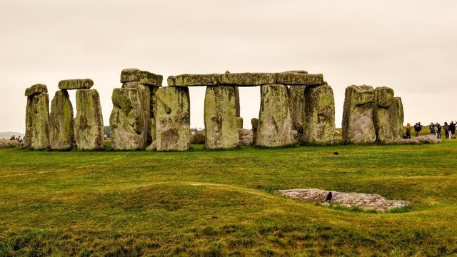 The image shows Stonehenge, a prehistoric monument located in Wiltshire, England. It features a ring of standing stones, each around 13 feet high, seven feet wide, and weighing approximately 25 tons. The stones are set within earthworks in the middle of the most dense complex of Neolithic and Bronze Age monuments in England. Stonehenge is widely believed to have been a ceremonial or religious site, possibly linked to astronomical events. The photograph captures the vastness and the mysterious atmosphere of the site, with a grassy landscape surrounding the stone circle. There are also visitors visible in the background, giving a sense of scale and the enduring popularity of this ancient site.
