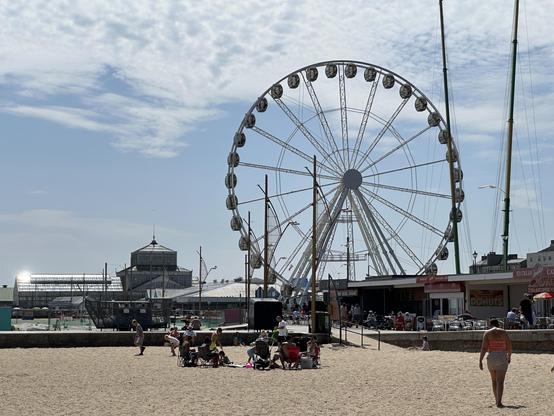 The ferris wheel at Great Yarmouth against a backdrop of blue sky and thin clouds on a hot, sunny day. The beach and various cafes are in the foreground, with lots of people enjoying the weather. In the background is the Victorian Winter Gardens, an enormous glasshouse, with sunlight glinting off one end.