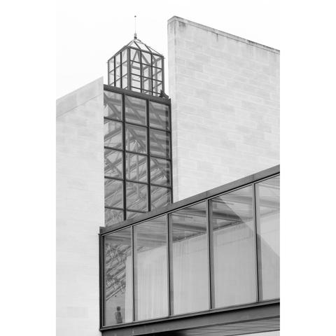 Black and white picture of a manin a lobby of the Mudam, in Luxembourg. The lobby is made of a succession of windows. The photography is taken from the outside. The other windows of the building and the small tower create the shape of an L.
