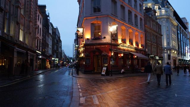 This image shows a picturesque street scene at dusk in an urban environment, possibly in a historic or older part of a city. Prominently displayed is a corner pub called "The Glassblower," which features bright warm lighting that contrasts with the twilight ambience of the street. The architecture has a traditional British style, suggesting the location might be in the UK, possibly London. The street is wet, reflecting the street lights and shop windows, indicating recent rain. There are some pedestrians visible, walking along the sidewalk or near the pub. The scene captures a classic, cozy urban vibe, typical of many British cities.