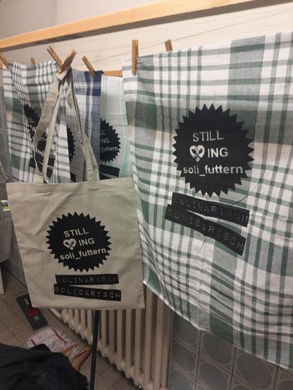 eine Wäscheleine mit diversen Küchentüchern und Jutebeutel zum trocknen, frisch bedruckt mit dem "Still <3i ng soli_futtern Logo" (Schrift im gezacktem Kreis) darunter 2 Balken mit der Schrift "kulinarisch solidarisch"
