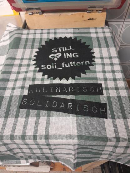 Küchentuch (weiss / dunklegrün kariert) efrisch bedruckt mit dem "Still <3i ng soli_futtern Logo" (Schrift im gezacktem Kreis) darunter 2 Balken mit der Schrift "kulinarisch solidarisch"  in schwarz