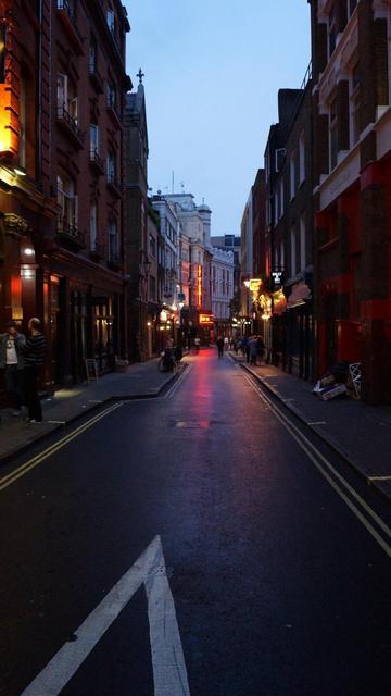 This image shows a narrow urban street scene at twilight. The street is lined with tall, mostly brick buildings that have a variety of businesses, including pubs and restaurants, evident from their illuminated signs. There's a noticeable red neon light further down the street contributing to the vibrant and lively atmosphere typical of a bustling city area. People are visible, some walking and others standing and chatting near the buildings. The road is wet, possibly from a recent rain, reflecting the lights and enhancing the evening mood of the setting. The setting gives an impression of a lively, possibly historic, city district, typical in many European cities.