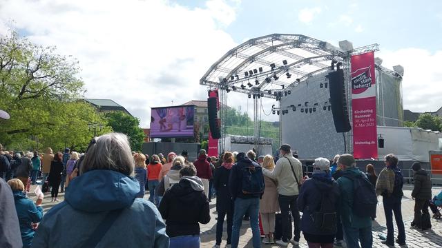 Menschen in bunter Kleidung vor einer Bühne und einer Videowand. Sonnenschein mit leichter Bewölkung.