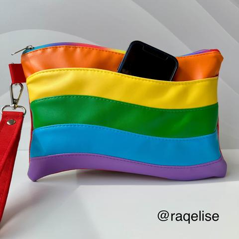 A hand sewn zippered pouch in the colors of the Pride flag. The shape is reminiscent of a flag waving. An iPhone sits in a side pocket of the pouch.