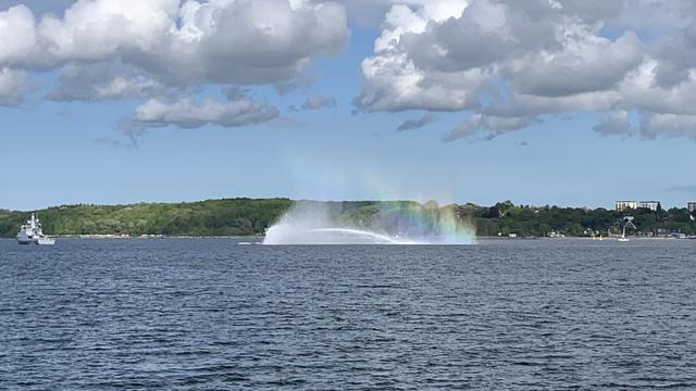 Das Bild zeigt eine malerische Szene auf einem ruhigen Wasser, vermutlich einem See oder einem Fluss. Im Vordergrund ist ein großes Wasserfontänensystem zu sehen, das einen prächtigen Regenbogen erzeugt. Der Regenbogen erstreckt sich über den Wasserstrahl und ist durch die Sonnenstrahlen und die feuchte Luft entstanden. Im Hintergrund sind bewaldete Ufer zu erkennen, die von einigen Gebäuden unterbrochen werden. Ein Schiff ist auf der linken Seite des Bildes zu sehen, das sich in der Ferne befindet. Der Himmel ist mit weißen, flauschigen Wolken bedeckt, die einen schönen Kontrast zum blauen Hintergrund bilden. Die gesamte Szene wirkt friedlich und idyllisch.
