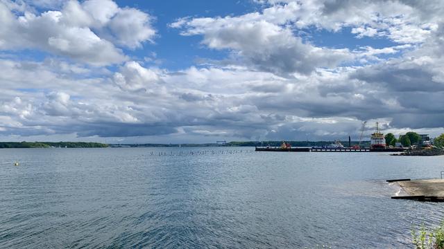 Das Bild zeigt eine ruhige Wasserlandschaft mit einem weiten Blick auf ein Gewässer, das von einer bewölkten Himmelsszene dominiert wird. Im Vordergrund erstreckt sich das Wasser, das von sanften Wellen gekräuselt wird. Auf der rechten Seite des Bildes ist ein Steg zu sehen, an dem ein Schiff und ein Baukran stehen, was auf eine maritime Aktivität hinweist. Im Hintergrund sind grüne Uferbereiche und ein weiterer Steg sichtbar, der sich in die Ferne erstreckt. Die Wolken am Himmel sind dicht und variieren in Größe und Form, was einen dynamischen Hintergrund bildet. Die Farben des Bildes sind natürlich und ruhig, mit einem dominierenden Blau des Wassers und des Himmels, kombiniert mit den verschiedenen Grautönen der Wolken.