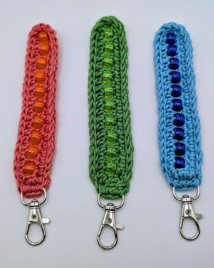 Three crochet fidget keychains; they are coral pink with orange pony beads, apple green with matching pony beads, and cerulean blue with dark blue pony beads.