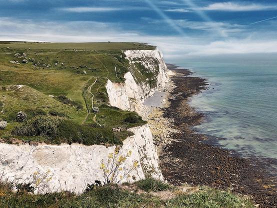This image shows a beautiful coastal scene featuring white chalk cliffs along a shoreline. The picture captures a vibrant blue ocean meeting with a rocky and pebbly beach. Towards the top of the cliffs, there's lush green grass, and a trail can be seen winding its way along the cliff side, likely a path for walkers. The sky is partly cloudy, suggesting a bright and pleasant day, which enhances the scenic beauty of the landscape. This type of landscape is typical of areas like the White Cliffs of Dover in England.