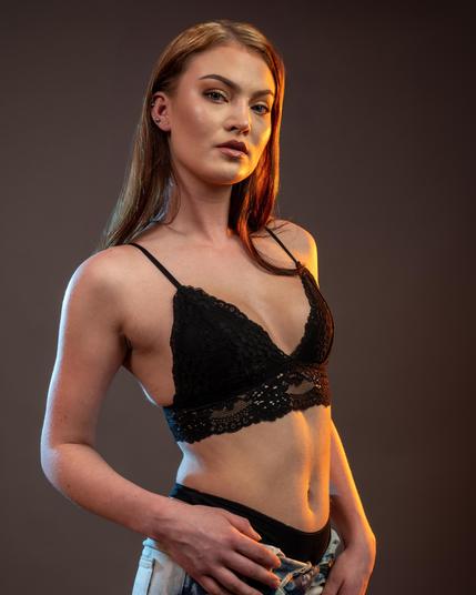 The image shows a person wearing a black lace bralette and holding a garment, possibly denim. The background is plain and the lighting is warm, highlighting their features. Valokuvaaja Lappeenranta Kuvaajankulma