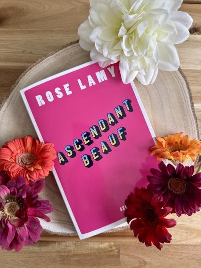 Livre posé sur un rondin de bois avec des fleurs autour. La couverture d’Ascendant beauf est rose vif avec le titre écrit en diagonale.