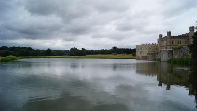 The image features a scenic view of a large body of water with a historic-looking castle on its edge. The castle has an old, robust architecture typical of medieval or early modern European structures, presented with stone walls and what appear to be defensive features. The sky is overcast, suggesting a cloudy day, and there is lush greenery in the background across the lake, enhancing the peaceful yet dramatic atmosphere of the setting. This looks like it could be a location in Europe, possibly the UK given the style of the castle and the landscape.