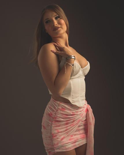 The image shows a person standing in a posed manner. They are wearing a white corset-style top and a pink and white patterned skirt. The person has long hair and is looking to the side with their hand resting on their chest. Valokuvaaja Lappeenranta Kuvaajankulma