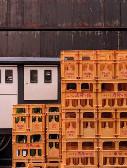 Bright orange boxes some filled with green glass bottles, others empty. They are located in front of the wooden wall of an old brewery.