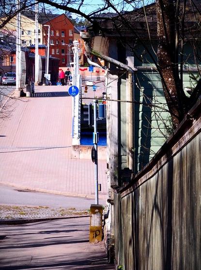 A view of a sloping street leading to a paved pathway. In the background, there are buildings and people walking, along with bicycle and pedestrian signs. Trees are in the foreground, and traffic signals are visible.