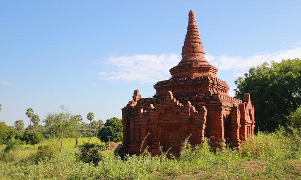 a very old stone and brick temple, which is low to the ground and small but still ornately decorated with spires, sits amid a grassy field filled with weeds, under a blue sky with light white clouds