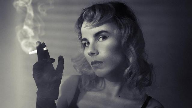 The image is in black and white and features a person with styled hair and makeup reminiscent of vintage fashion. They are wearing a glove and holding a cigarette, with smoke visible in the air. The lighting and composition give it a classic, possibly film noir aesthetic. Valokuvaaja Kuvaajankulma Lappeenranta