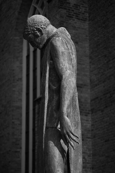 A monochrome image of a textured bronze statue of St George, with its head bowed, draped in a robe, standing beside a brick wall. The pose conveys a sense of contemplation or sorrow.