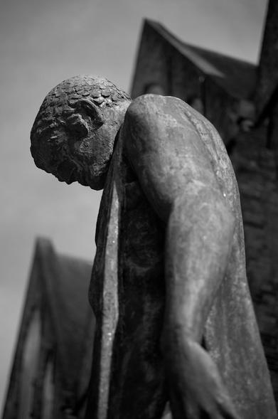 A close-up of a weathered bronze statue of a man with his head bowed, featuring intricate details on his face and body. The background includes blurred elements of a building, captured in black and white.