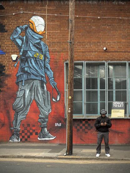 A street scene with a red brick building. On the building is a street art sci-fi esk figure wearing a helmet. In front of the wall a man is looking at his phone while wearing headphones. Perhaps the street art figure is looking at  the man, but it's difficult to tell as they are depicted wearing a helmet.