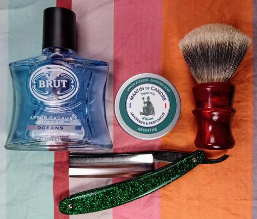 (Le Chat Mistral description)

This image shows a collection of men's grooming items arranged on a striped fabric background. On the left, there's a blue glass bottle of Brut aftershave labelled "Océans". Next to it is a round tin of shaving soap by Martin de Candre, featuring an absinthe scent. To the right, there's a red-handled shaving brush with a fluffy top. Below these items, a straight razor with a green glittery handle is placed. The overall scene is vibrant and colourful, highlighting classic shaving essentials.