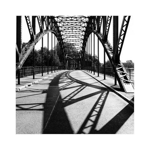 Brücke mit beidseitigen Stahlbögen, welche oberhalb mit Traversen verbunden sind. Auf dem Asphalt der Schattenwurf des rechten Bogens und seiner Streben
