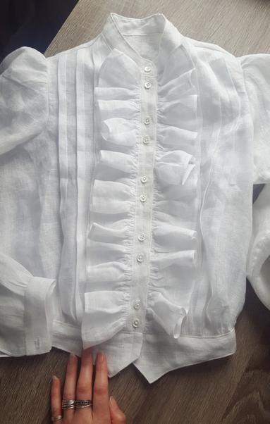 A top down photo of a white linen blouse in an Edwardian style, with tucks and lovely full ruffles. It's from an airy and lightweight linen, sheer and soft. There are 10 tiny shell buttons with four holes along a narrow button placket, in pairs of two. They are small and pearlescent. Sini's hand in lower frame, blouse looking done here but still missing a couple of things not visible in this photo.
