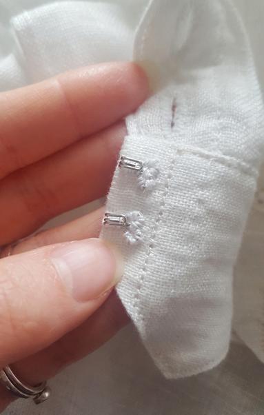 Close up photo of the tiny hooks portion of the hooks and bars closures on the shaped waistband. They are very small and the bit that attaches to the garment has been nearly fully covered in tiny stitches of white linen thread. Two of the buttonholes are visible from the wrong side. They have been neatly sewn with the same linen thread, well contained, oval and small.