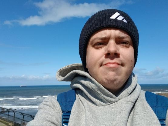 A white transmasc wearing a black beanie, a grey hoodie, and a blue rucksack. He's stood in front of the north sea.