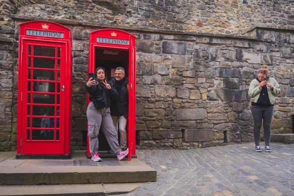 A vibrant photo taken at Edinburgh Castle in Scotland, showing two people posing for a selfie in front of classic red British telephone boxes, with another person standing nearby, all set against the historic stone walls of the castle; the word "TELEPHONE" is clearly visible atop both phone boxes.