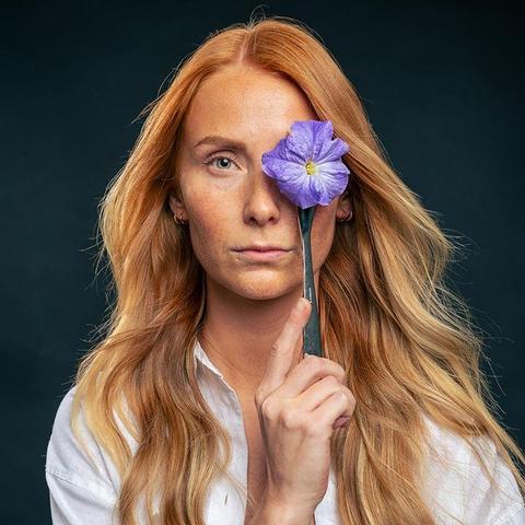 The image shows a person with long, reddish-blonde hair holding a purple flower in front of one eye. The background is dark, and the person appears to be wearing a white shirt. Valokuvaaja Kuvaajankulma Lappeenranta