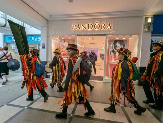 Powderkegs walking through an indoor shopping centre, in front of the Pandora shop. They wear jackets with long tatters, strips of fabric in red, orange, yellow and black, over black shirts and trousers. They wear bell pads on their legs with short tatters, and black top hats decorated with different items including ribbons, flowers and feathers. Their faces are painted in designs in the same colours as their jackets.