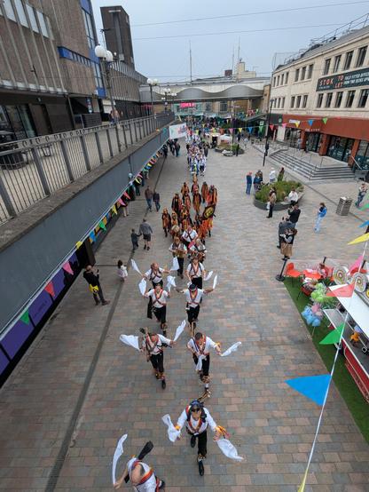 Morris dancers parading through a town centre street lined with shops. Cotswold dancers in white shirts and colourful baldricks wave white hankies as they dance. Powderkegs walk behind.