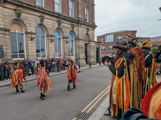 Four Powderkegs dancers in the road, two throw their sticks to each other and the other two tap the bottom of their sticks on the floor. Three musicians stand to the right on the pavement. On the opposite side, a line of people stand watching.