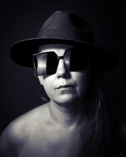 The image is a black and white portrait of a person wearing a fedora hat and large square sunglasses. They appear to have long hair, and the expression is neutral. The background is dark, highlighting the person's face and accessories. Valokuvaaja Lappeenranta Kuvaajankulma